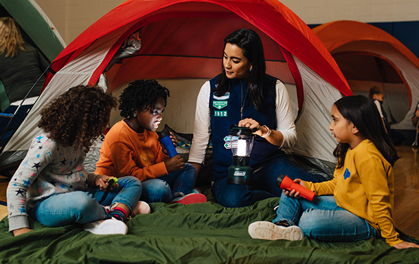 a girl scout volunteer sitting in front of a tent with a group of girl scouts telling stories by lantern and flashlight