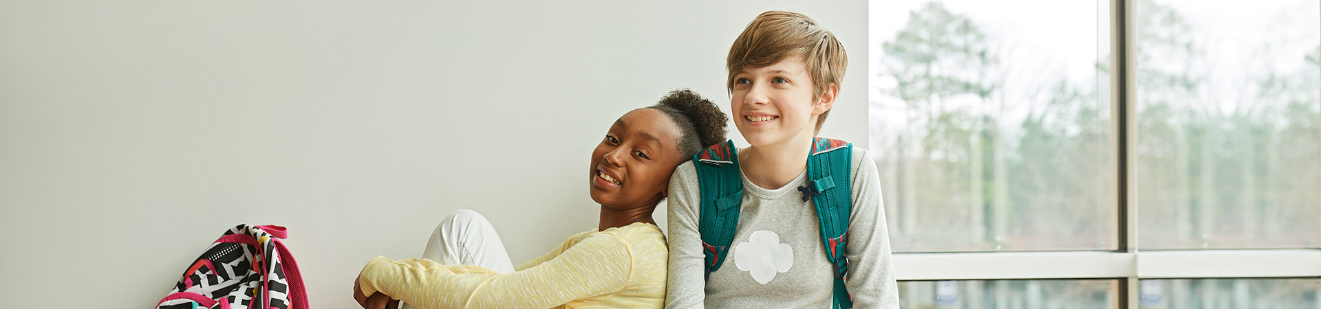  two girl scouts wearing trefoil clothing leaning on one another indoors 