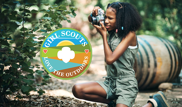 Photo of a Girl Scout kneeling and taking a photo outdoors with an image of a patch featuring the text Girl Scouts Love the Outdoors